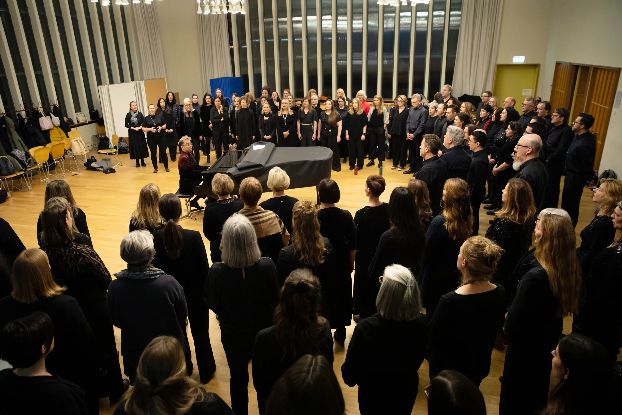 Grosser Chor in schwarzer Kleidung steht im Kreis um einen Flügel in einem hellen Saal mit Parkettboden