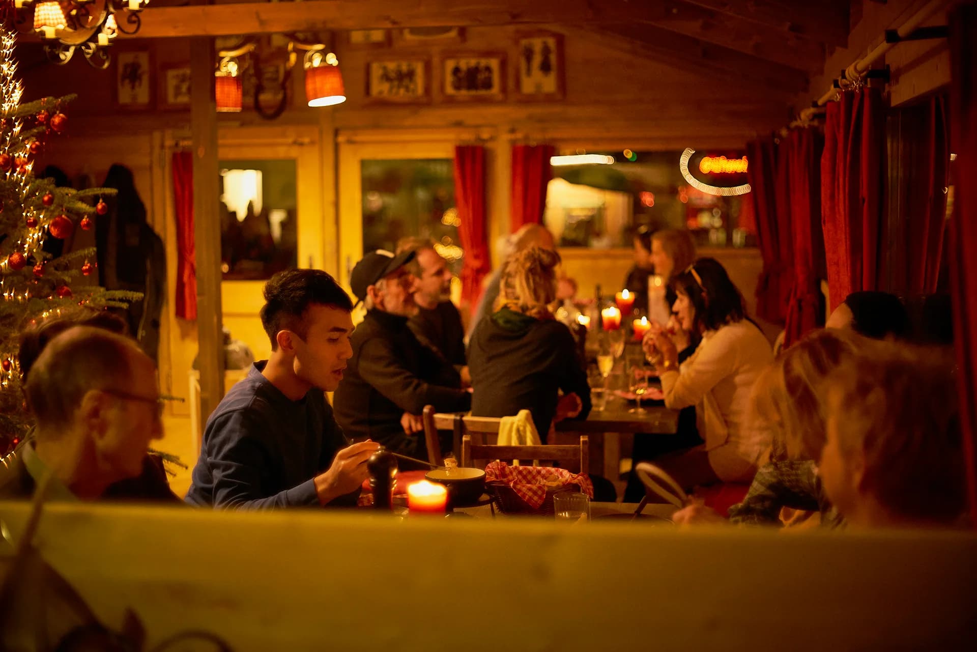 Gäste sitzen im Fondue Chalet beim Abendessen, Kerzen auf den Tischen und ein Tannenbaum im Hintergrund