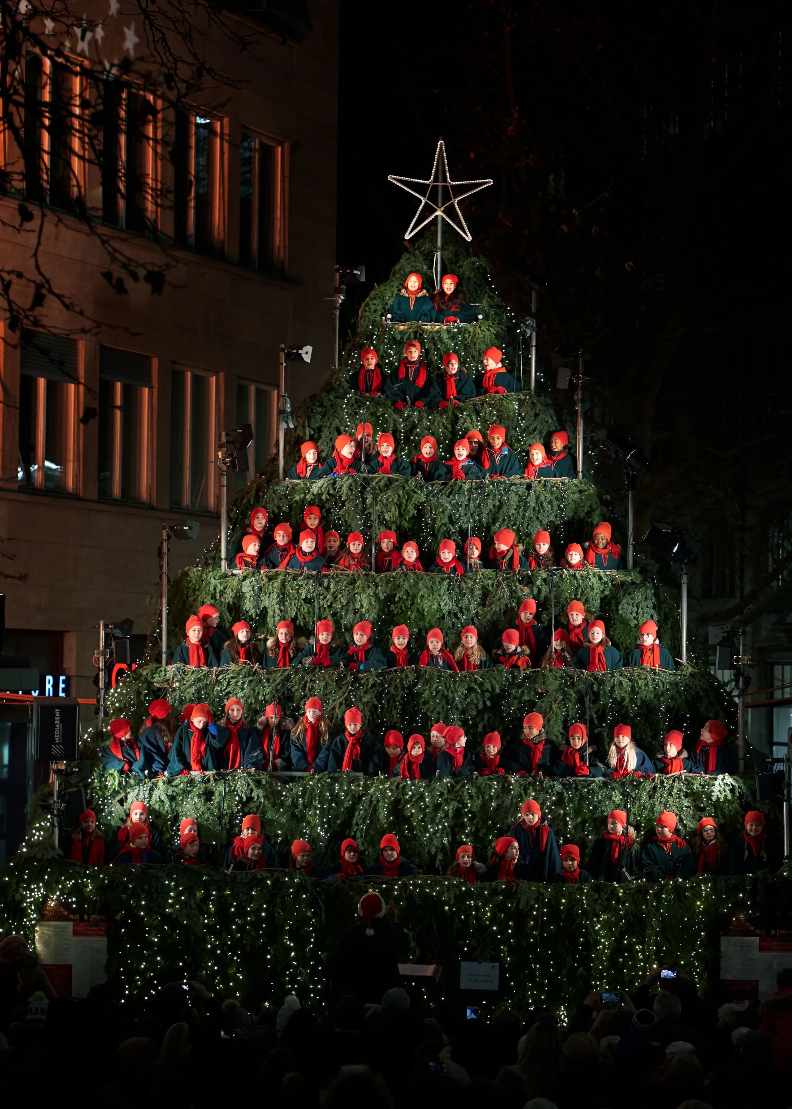 Coro di bambini con berretti e sciarpe rossi sui gradini del Singing Christmas Tree illuminato di notte