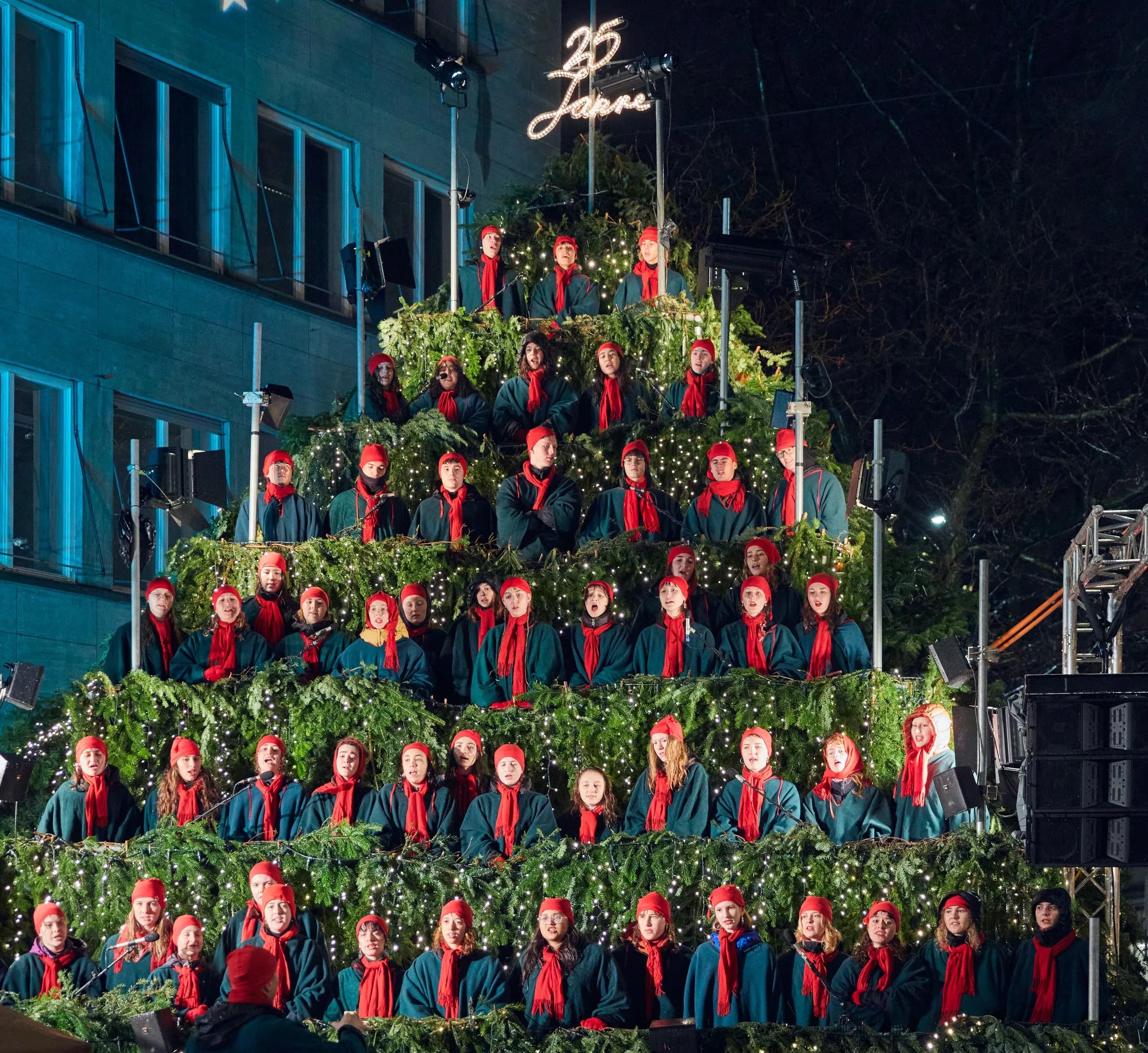 Il Singing Christmas Tree con la scritta luminosa «25 Jahre»: coro con berretti e sciarpe rossi sui gradini del palco