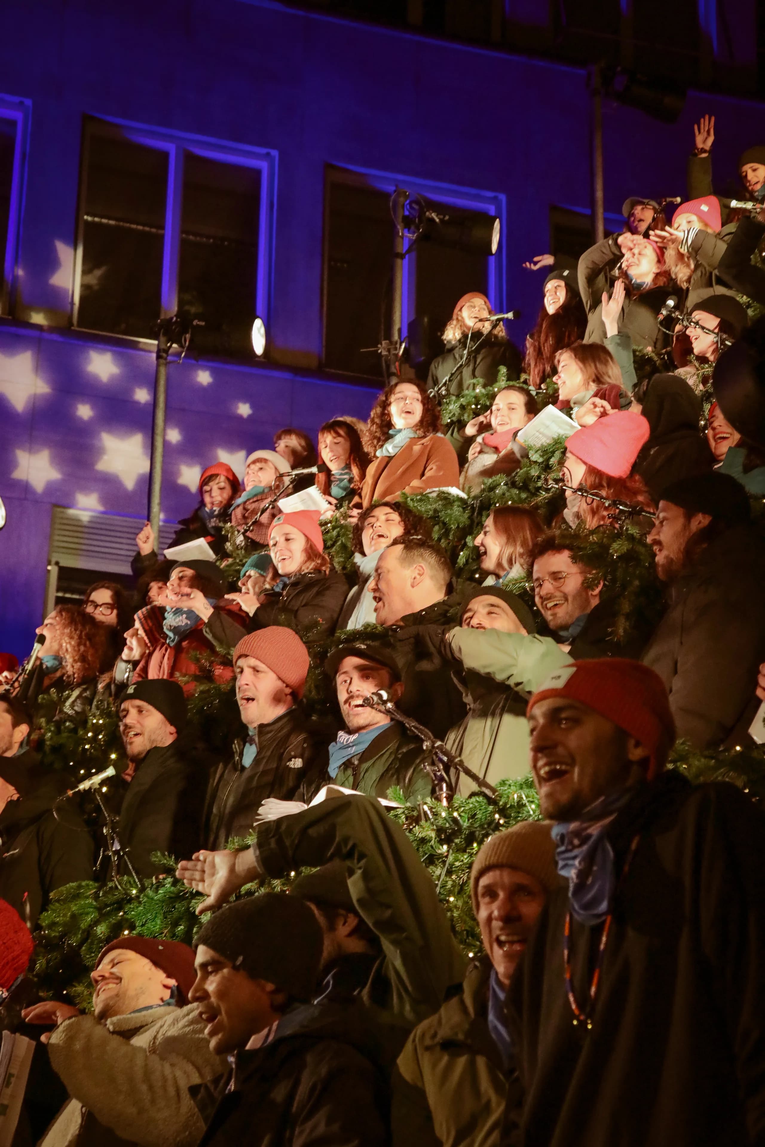 Offenes Singen am Abend: Menschen in Winterkleidung singen auf der Baumbühne vor einer blau beleuchteten Fassade mit Sternen