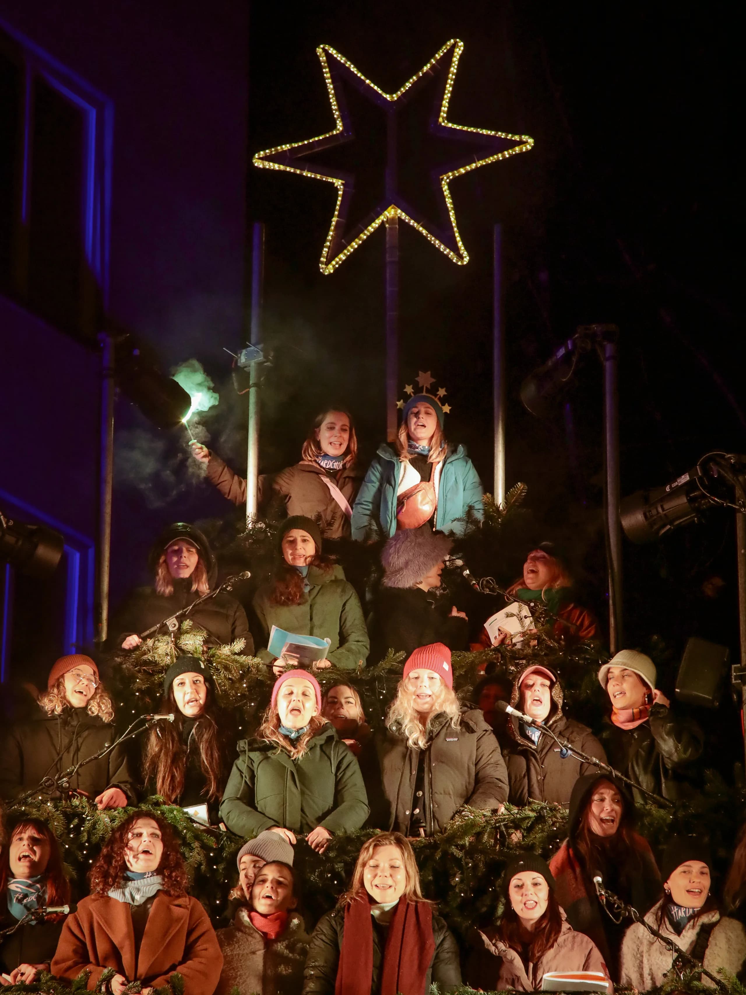 Persone che cantano sui gradini più alti del Singing Christmas Tree sotto la stella di luci, una alza una luce