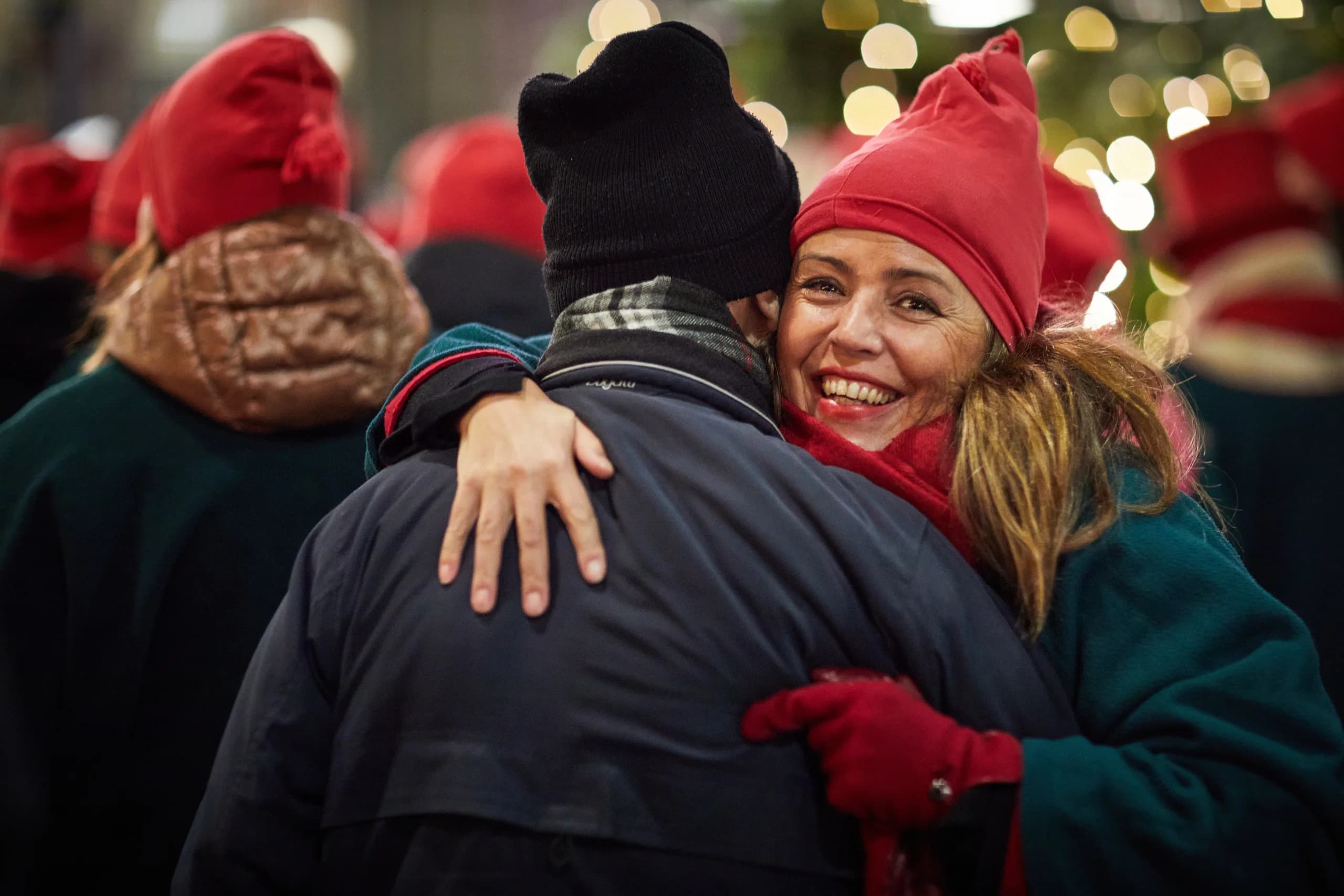 Frau mit roter Mütze und roten Handschuhen umarmt lachend einen Mann mit dunkler Mütze, dahinter weihnachtliche Lichter