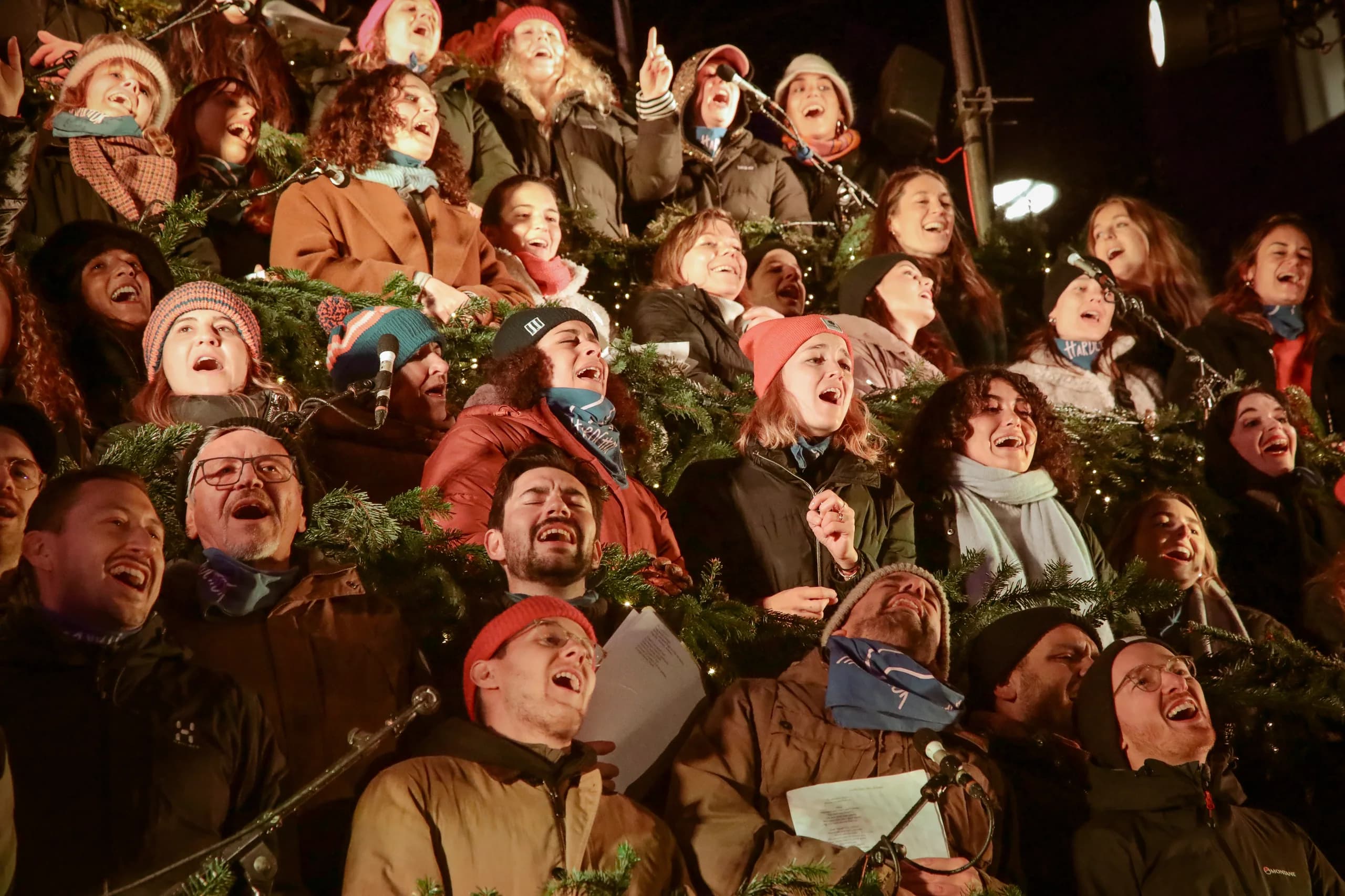 Molte persone in giacche invernali cantano e ridono insieme sui gradini del Singing Christmas Tree durante il canto aperto