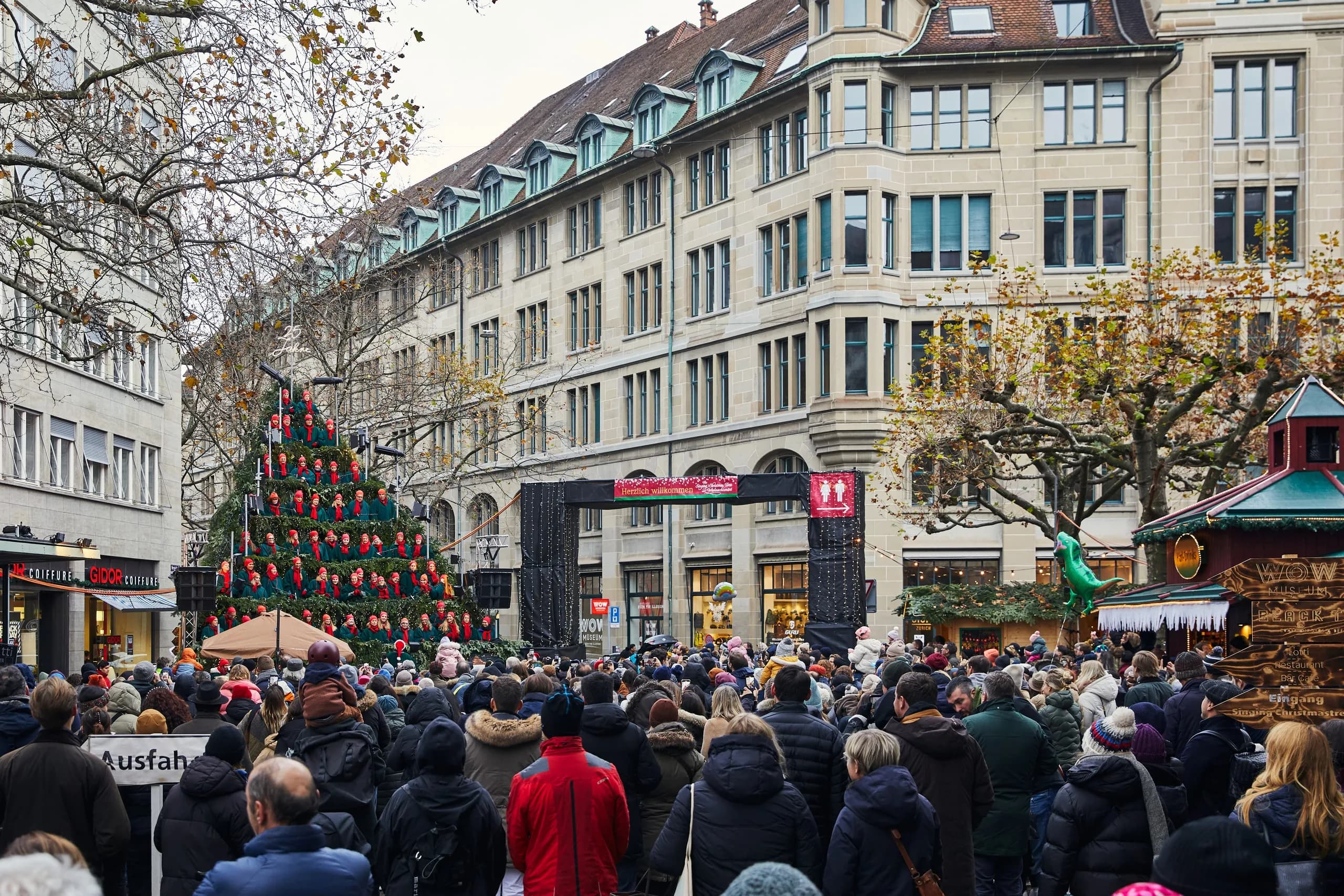 Large crowd in winter jackets on Werdmühleplatz by day, facing the Singing Christmas Tree with the illuminated sign «25 Jahre», next to it the entrance arch to the Christmas market
