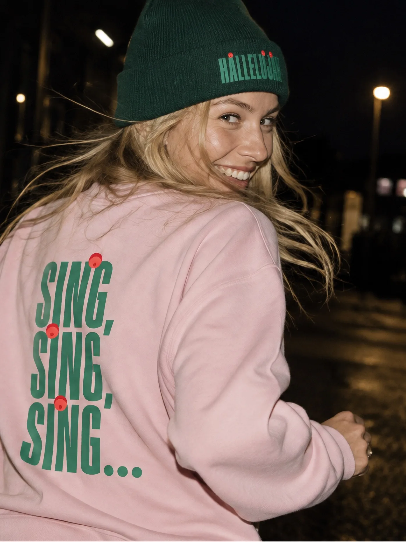 Lachende Frau mit grüner «Halleluja»-Mütze und rosa Pulli mit dem Aufdruck «Sing, Sing, Sing…» – Merchandise des Singing Christmas Tree