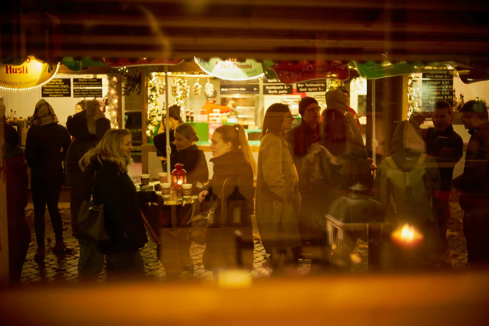 Besucherinnen und Besucher an Stehtischen vor den beleuchteten Markthäuschen am Wiehnachtsmärt, durch eine Scheibe gesehen
