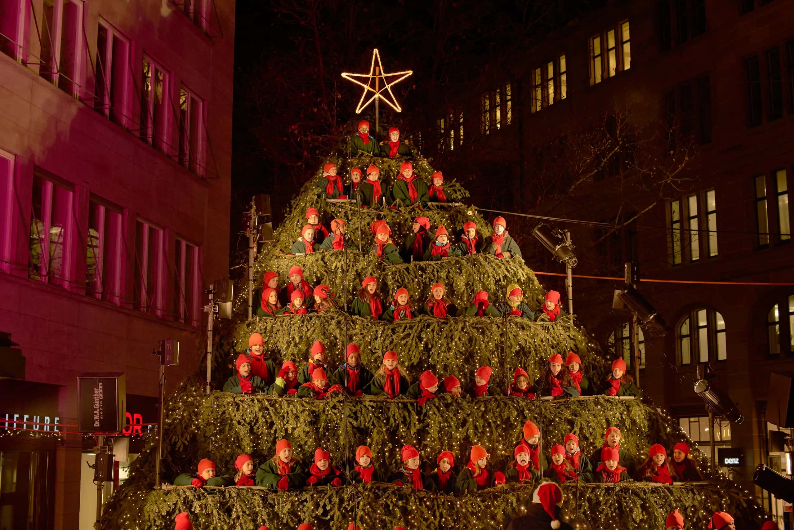 Der Singing Christmas Tree am Werdmühleplatz Zürich bei Nacht: Kinderchor in Rot auf der Baumbühne unter einem Lichterstern