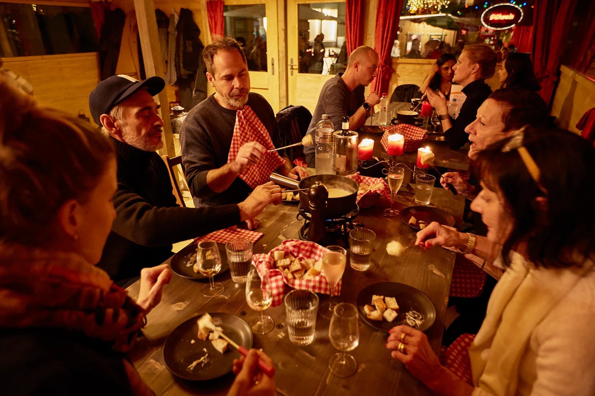 Guests eating cheese fondue at a long wooden table in the fondue chalet, with candles, wine glasses and bread baskets