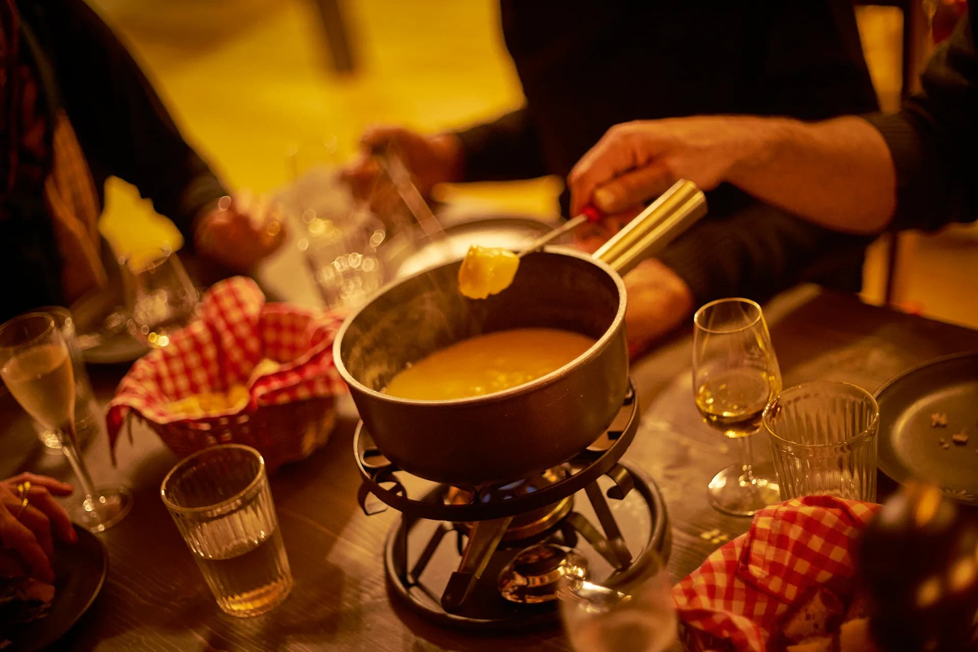 Brotstück an der Fonduegabel über dem Caquelon mit Käsefondue auf dem Rechaud, daneben Weissweingläser und Brotkorb