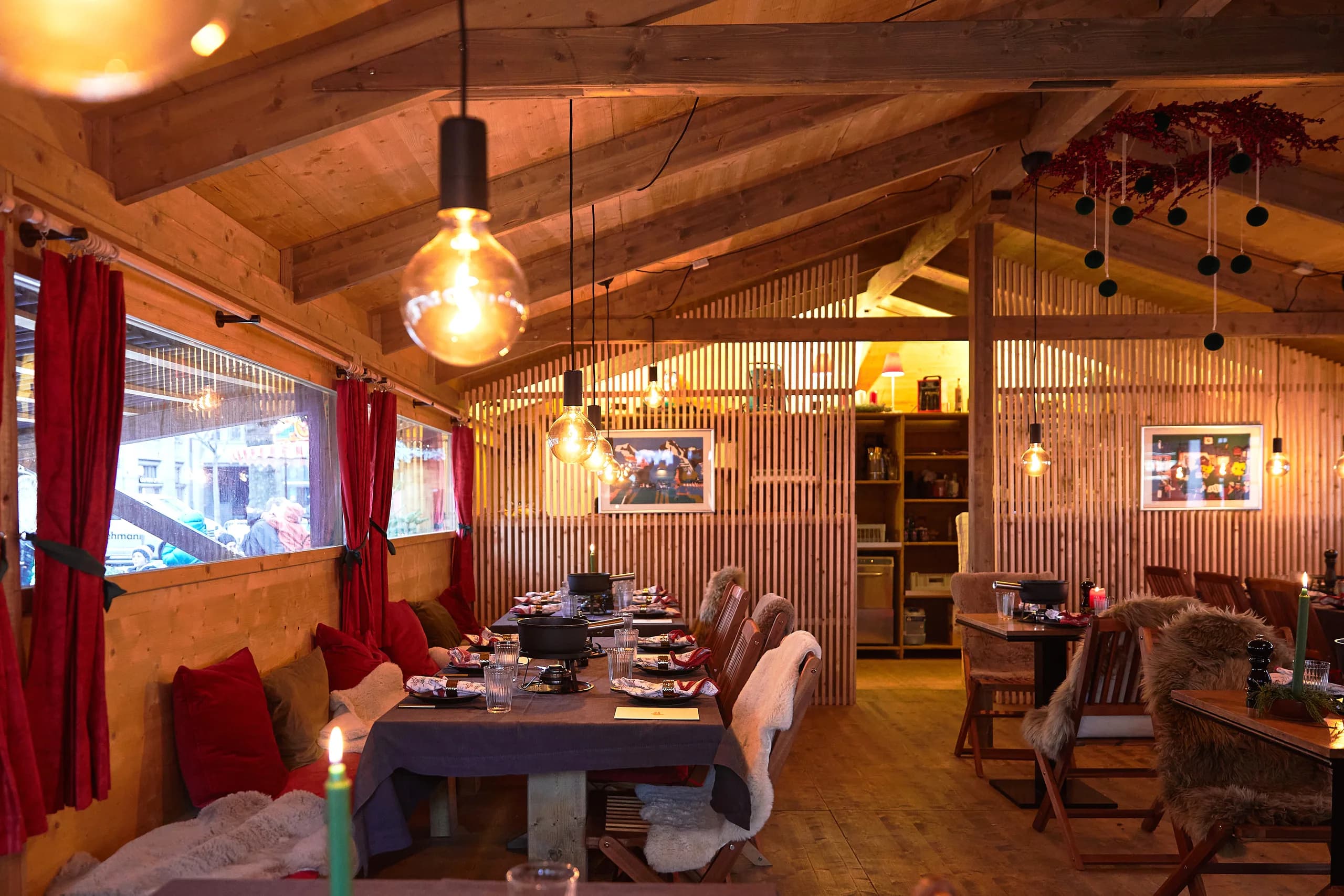 Laid tables in the wood-panelled fondue chalet with hanging light bulbs, red cushions, furs and windows onto the market