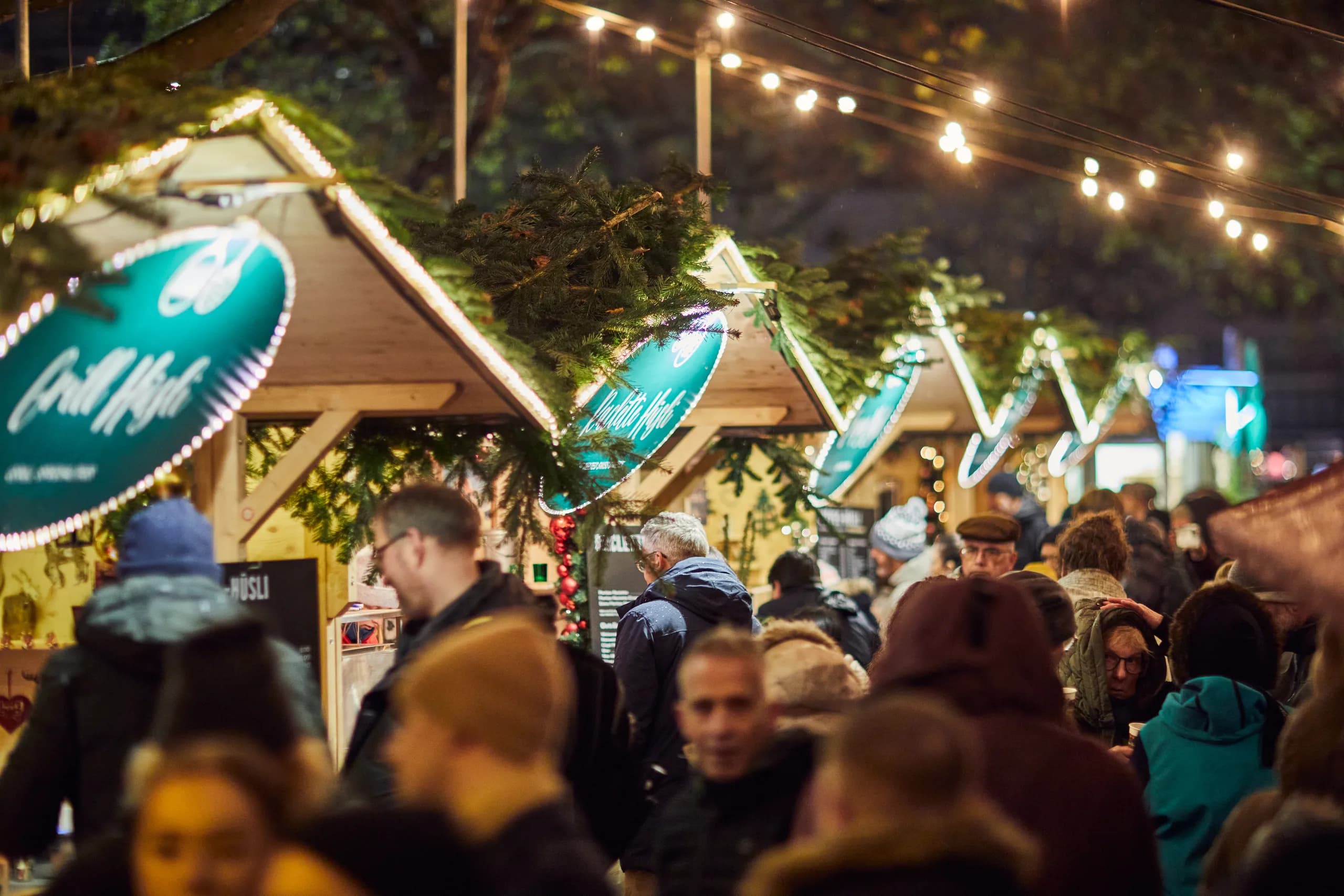 Illuminated market huts with fir branches and fairy lights at the Christmas market, visitors in front of them in the evening
