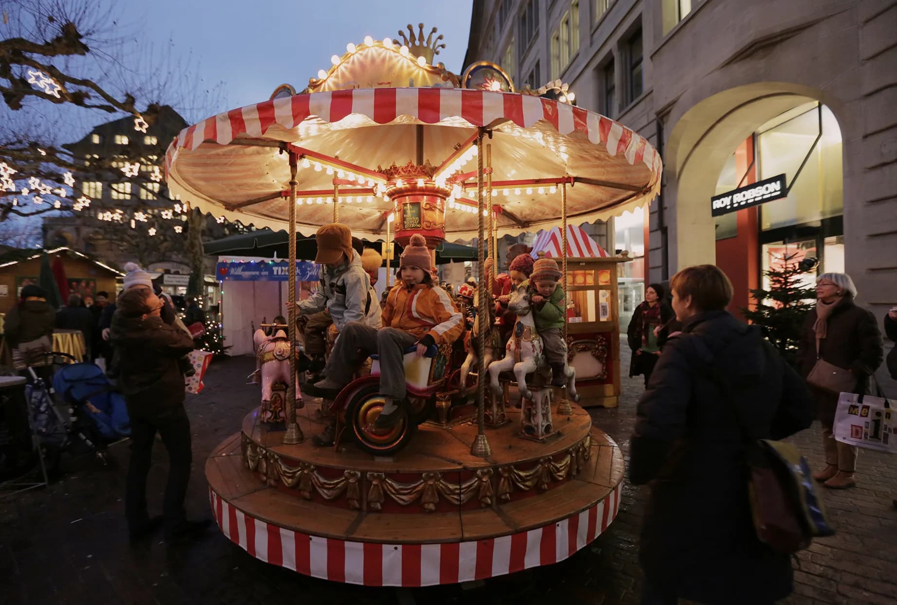 Children riding the red-and-white striped, illuminated children's carousel at the Christmas market at dusk