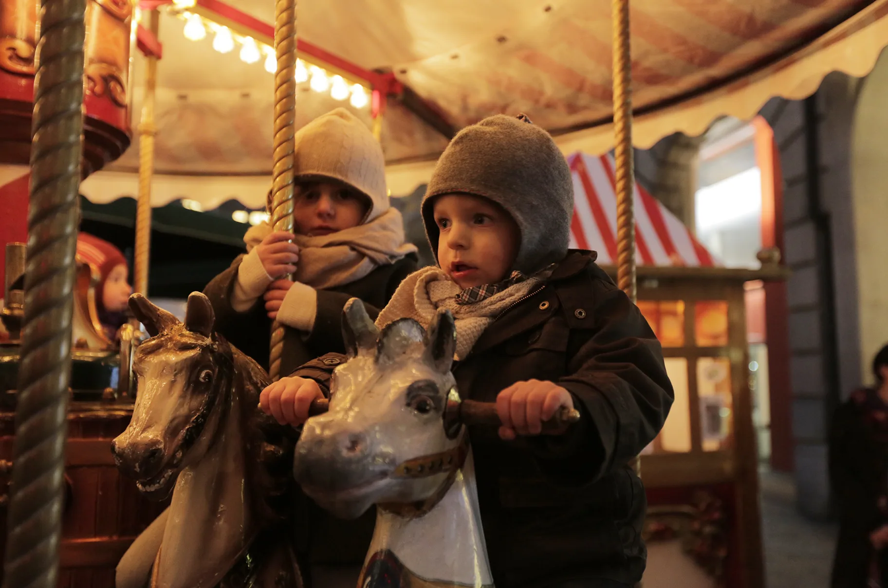 Two small children in woolly hats and scarves riding painted wooden horses on the children's carousel