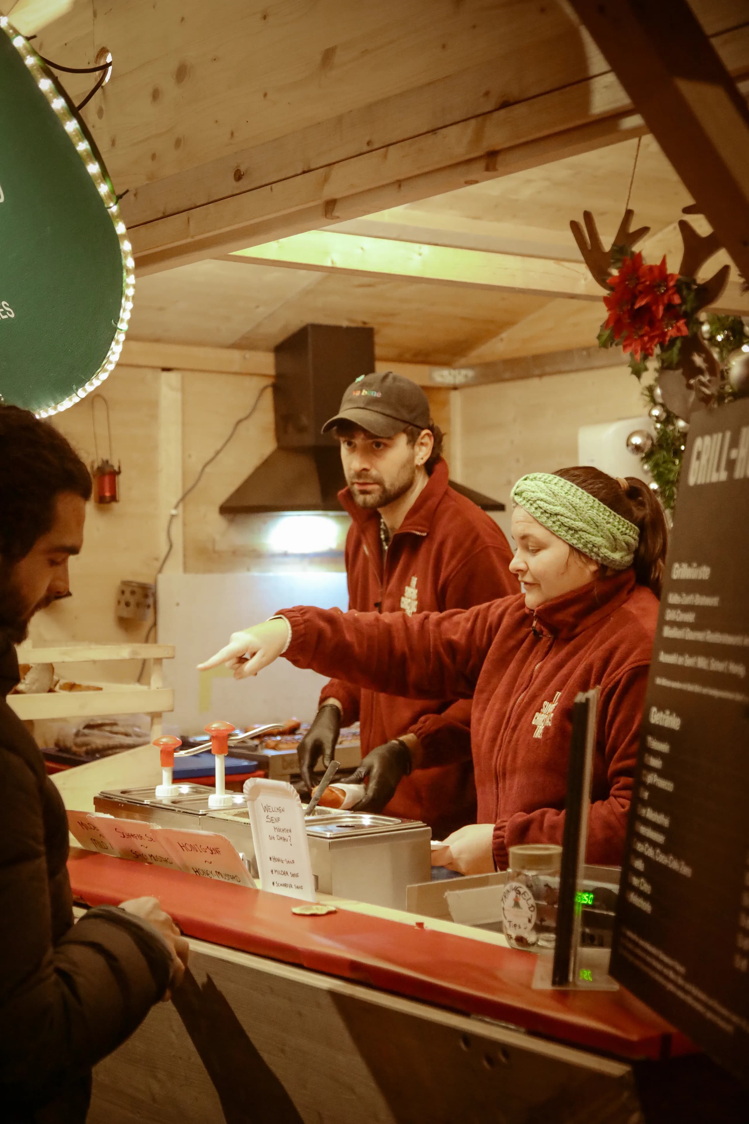 Two staff members in red fleece jackets serving a guest at the grill stand of a Christmas market hut